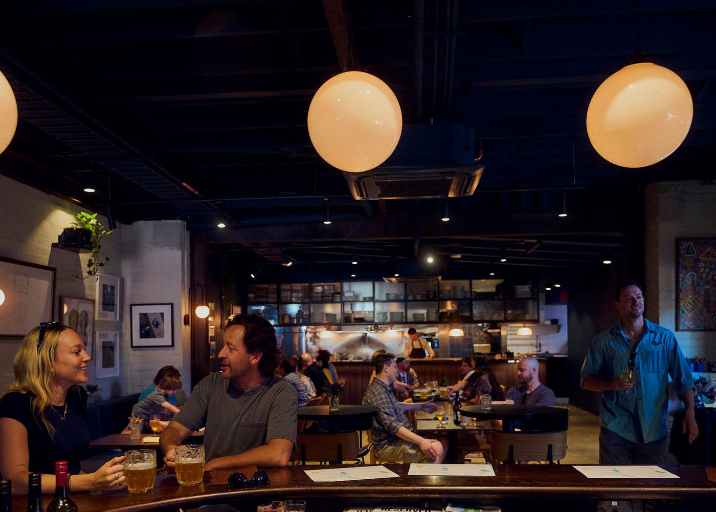 Dining area with people seated at tables and a man standing, illuminated by large spherical lights.